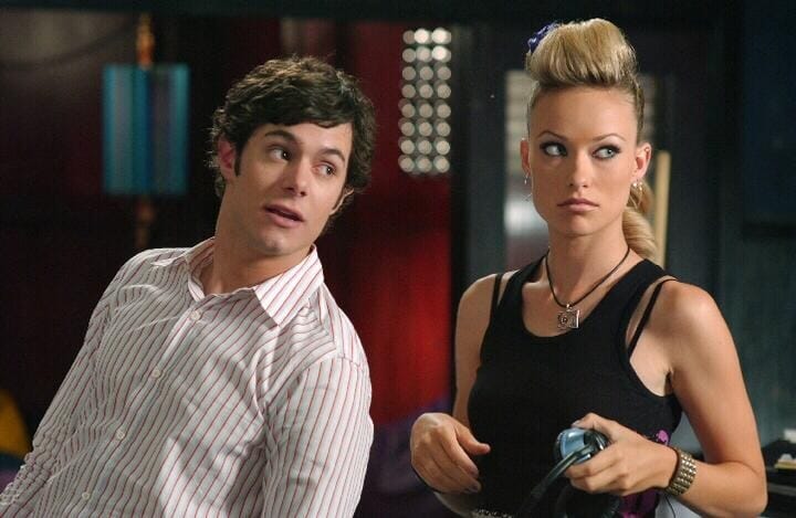 Adam Brody wears a striped button up and tilts toward Olivia Wilde in a black tank top with lots of jewelry at the Bait Shop