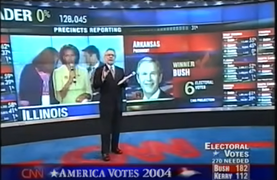 Wolf Blitzer stands in CNN's 2004 election night control room. Michelle Obama is  on a screen behind him and he ignores her.