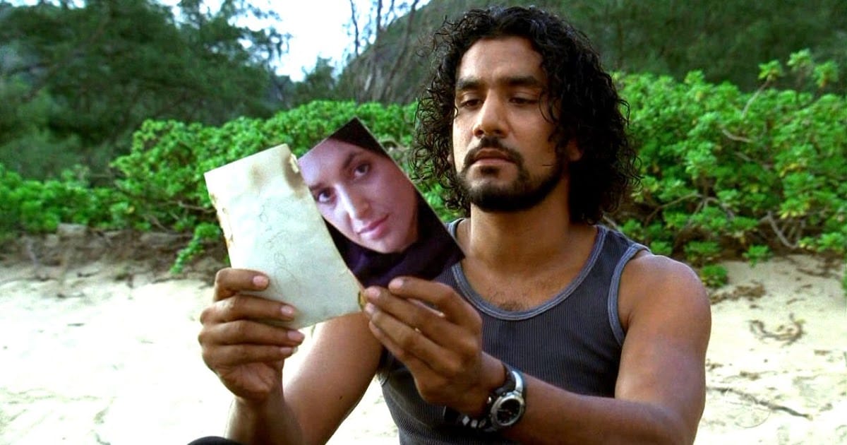 Sayid sits on the beach in a black tank top and looks at a photo of a woman. Green bushes in the background.