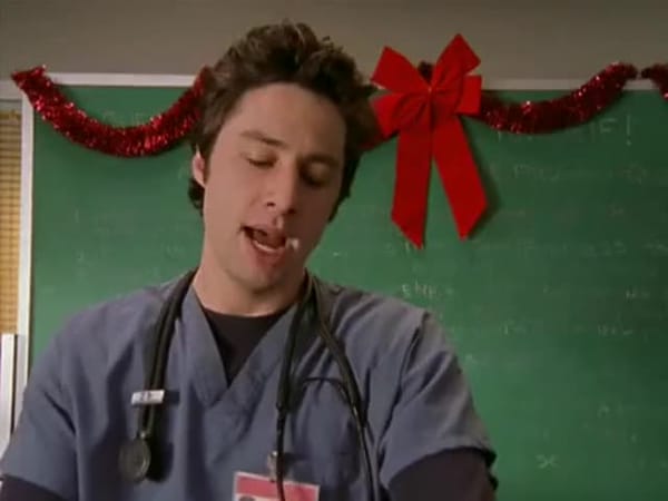 JD stands in front on a chalkboard decorated with Xmas garland and bow, wearing scrubs and stethoscope